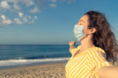 Mavi gökyüzü arka planında tıbbi maske takan mutlu bir kadın. Yazın deniz kenarında eğlenen kişi. Coronavirus salgını ve sağlıklı yaşam tarzı konsepti