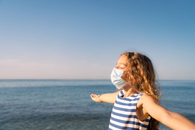 Mavi gökyüzü arka planında tıbbi maske takan mutlu bir çocuk. Kız yazın deniz kenarında eğleniyor. Coronavirus salgını ve sağlıklı yaşam tarzı konsepti