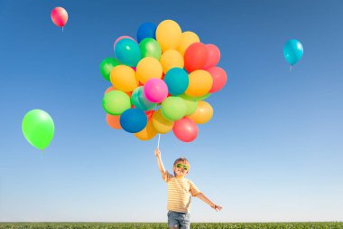 Mutlu çocuk açık havada parlak renkli balonlarla oynuyor. Çocuk mavi gökyüzü arka planına karşı yeşil bahar tarlasında eğleniyor. Sağlıklı ve aktif yaşam tarzı kavramı