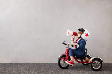 Gri beton arka planda bisiklet süren takım elbiseli mutlu çocuk. Hoparlörden bağıran komik çocuk. Çocukluk hayalleri ve iş fikri