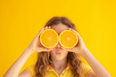 Çocuk güneş gözlüğü yerine turuncu yarımlar tutuyor. Sarı arka planda şaşırmış bir kızın portresi
