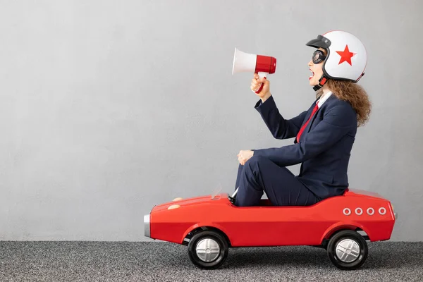 Successfull businesswoman driving toy car outdoor. Funny young woman shouting through megaphone against concrete wall background. Business srart up and winner concept