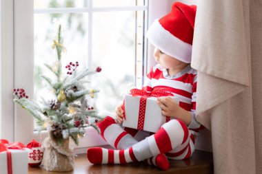 Pencerenin kenarında oturan çocuk. Mutlu çocuk Noel pijaması giyiyor. Elinde Noel hediyesi olan bir çocuk. Gülümseyen çocuğun portresi