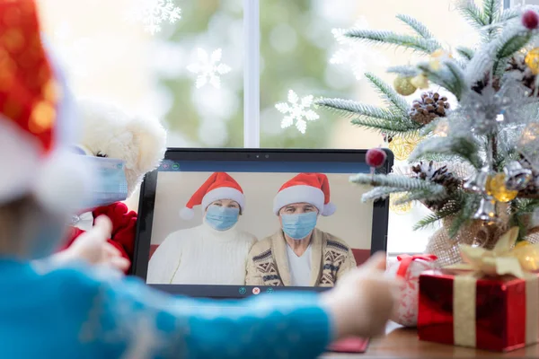 医者のマスクをした幸せな子供 おじいちゃんとおばあちゃんのビデオチャットで挨拶 クリスマスツリーの装飾に対する窓辺のラップトップ コロナウイルスのCovid 19概念中のクリスマス休暇 家にいて — ストック写真