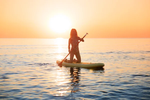 Genç bir kadın yaz tatilinde deniz kenarında ayağa kalkıp kürek çekiyor. Güzel bir kadın deniz ve gökyüzüne karşı kürek çekiyor.