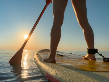 Genç bir kadın yaz tatilinde deniz kenarında ayağa kalkıp kürek çekiyor. Güzel bir kadın deniz ve gökyüzüne karşı kürek çekiyor.