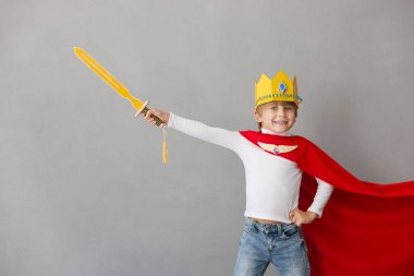 Komik çocuk şövalye gibi davranıyor. Mutlu çocuk evde eğleniyor. Kırmızı pelerin ve gri arka planda kağıt kaplama giyen küçük bir çocuk. Çocukların hayal gücü ve özgürlük kavramı