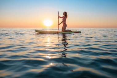 Genç bir kadın yaz tatilinde deniz kenarında ayağa kalkıp kürek çekiyor. Güzel bir kadın deniz ve gökyüzüne karşı kürek çekiyor.