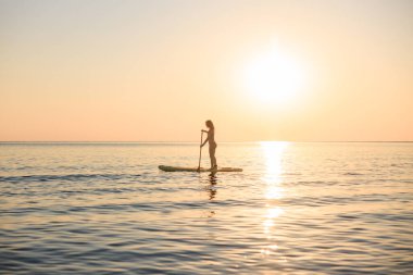 Genç bir kadın yaz tatilinde deniz kenarında ayağa kalkıp kürek çekiyor. Güzel bir kadın deniz ve gökyüzüne karşı kürek çekiyor.