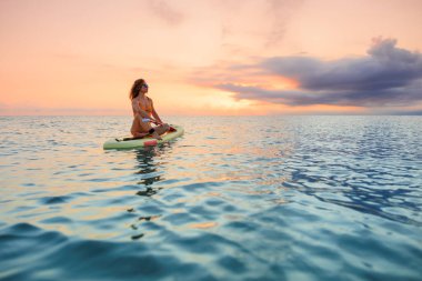 Genç bir kadın yaz tatilinde deniz kenarında ayağa kalkıp kürek çekiyor. Deniz ve gökyüzü arka planında kürek çeken güzel bir kız.