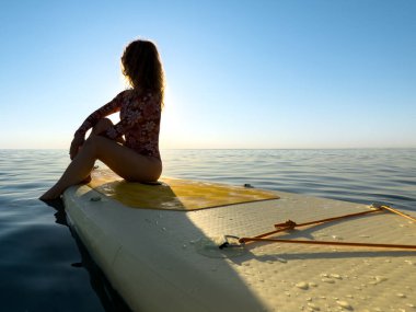 Genç bir kadın yaz tatilinde deniz kenarında ayağa kalkıp kürek çekiyor. Güzel bir kadın deniz ve gökyüzüne karşı kürek çekiyor.