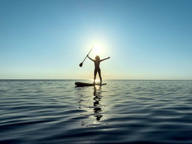 Genç bir kadın yaz tatilinde deniz kenarında ayağa kalkıp kürek çekiyor. Güzel bir kadın deniz ve gökyüzüne karşı kürek çekiyor.
