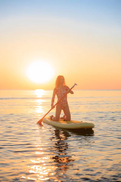 Genç bir kadın yaz tatilinde deniz kenarında ayağa kalkıp kürek çekiyor. Güzel bir kadın deniz ve gökyüzüne karşı kürek çekiyor.