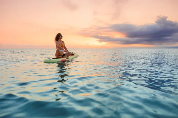 Genç bir kadın yaz tatilinde deniz kenarında ayağa kalkıp kürek çekiyor. Deniz ve gökyüzü arka planında kürek çeken güzel bir kız.