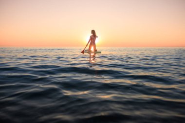 Genç bir kadın yaz tatilinde deniz kenarında ayağa kalkıp kürek çekiyor. Güzel bir kadın deniz ve gökyüzüne karşı kürek çekiyor.