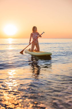 Genç bir kadın yaz tatilinde deniz kenarında ayağa kalkıp kürek çekiyor. Güzel bir kadın deniz ve gökyüzüne karşı kürek çekiyor.
