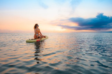 Genç bir kadın yaz tatilinde deniz kenarında ayağa kalkıp kürek çekiyor. Deniz ve gökyüzü arka planında kürek çeken güzel bir kız.