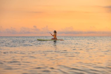 Genç bir kadın yaz tatilinde deniz kenarında ayağa kalkıp kürek çekiyor. Deniz ve gökyüzü arka planında kürek çeken güzel bir kız.
