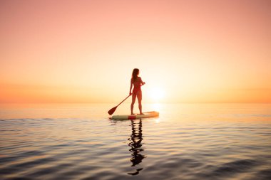 Genç bir kadın yaz tatilinde deniz kenarında ayağa kalkıp kürek çekiyor. Deniz ve gökyüzü arka planında kürek çeken güzel bir kız.