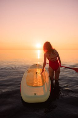 Genç bir kadın yaz tatilinde deniz kenarında ayağa kalkıp kürek çekiyor. Deniz ve gökyüzü arka planında kürek çeken güzel bir kız.