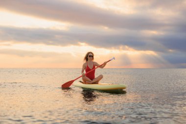 Genç bir kadın yaz tatilinde deniz kenarında ayağa kalkıp kürek çekiyor. Deniz ve gökyüzü arka planında kürek çeken güzel bir kız.