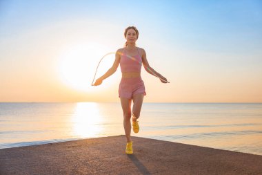 Sahilde deniz ve gökyüzü arka planına karşı ip atlayan genç bir kadın. Yaz sporu ve sağlıklı yaşam tarzı konsepti