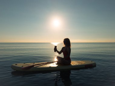 Genç bir kadın yaz tatilinde deniz kenarında ayağa kalkıp kürek çekiyor. Güzel kız suda yüzüyor ve deniz ve gökyüzü arka planına karşı kahve içiyor.
