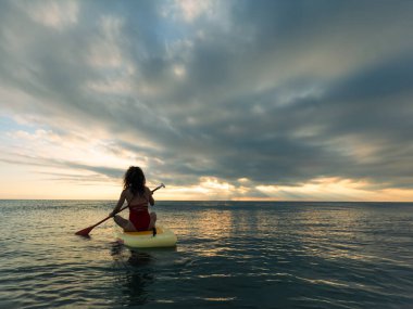 Genç bir kadın yaz tatilinde deniz kenarında ayağa kalkıp kürek çekiyor. Deniz ve gökyüzü arka planında kürek çeken güzel bir kız.
