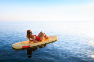 Genç bir kadın yaz tatilinde deniz kenarında ayağa kalkıp kürek çekiyor. Deniz ve gökyüzü arka planında kürek çeken güzel bir kız.