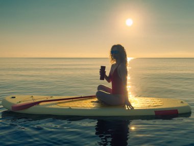 Genç bir kadın yaz tatilinde deniz kenarında ayağa kalkıp kürek çekiyor. Güzel kız suda yüzüyor ve deniz ve gökyüzü arka planına karşı kahve içiyor.