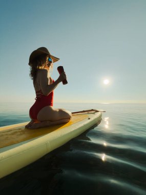 Genç bir kadın yaz tatilinde deniz kenarında ayağa kalkıp kürek çekiyor. Güzel kız suda yüzüyor ve deniz ve gökyüzü arka planına karşı kahve içiyor.