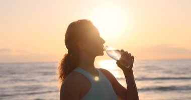 Sabahları açık havada koştuktan sonra sahilde su içen zinde bir kadın. Sportif kadın antrenmandan sonra dinleniyor. Yaz sporları ve sağlıklı yaşam tarzı konsepti. Yavaş çekim
