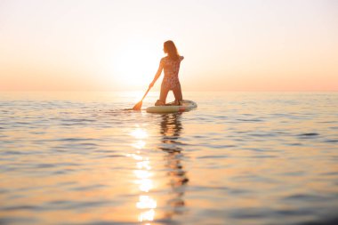 Genç bir kadın yaz tatilinde deniz kenarında ayağa kalkıp kürek çekiyor. Güzel bir kadın deniz ve gökyüzüne karşı kürek çekiyor.