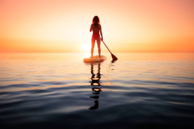 Yaz tatili boyunca denizin kenarında ayakta kürek çeken genç bir kadının silueti. Güzel bir kadın denize ve gündoğumuna karşı kürek çekiyor. Sağlıklı yaşam tarzı kavramı