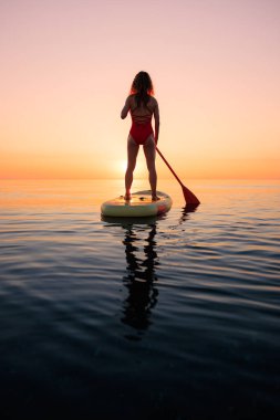 Yaz tatili boyunca denizin kenarında ayakta kürek çeken genç bir kadının silueti. Güzel bir kadın denize ve gündoğumuna karşı kürek çekiyor. Sağlıklı yaşam tarzı kavramı