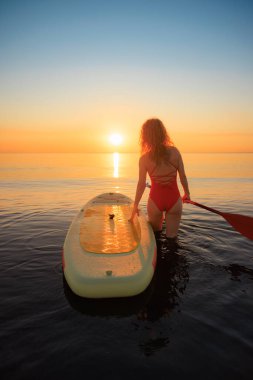 Genç bir kadın yaz tatilinde denize karşı kürek çekerek ayakta yürüyor. Güzel bir kadın denize ve gündoğumuna karşı kürek çekiyor. Sağlıklı yaşam tarzı kavramı