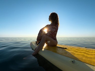Genç bir kadın yaz tatilinde deniz kenarında ayağa kalkıp kürek çekiyor. Güzel bir kadın deniz ve gökyüzüne karşı kürek çekiyor.