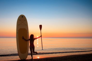 Yaz tatili boyunca ayakta ayakta duran genç bir kadın, deniz kenarında kürek çekiyor. Güzel kız denizlere ve gökyüzüne karşı