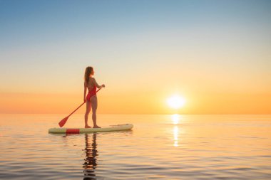 Genç bir kadın yaz tatilinde deniz kenarında ayağa kalkıp kürek çekiyor. Deniz ve gökyüzü arka planına karşı kürek çeken güzel bir kız. Sağlıklı yaşam tarzı kavramı