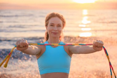Sahilde deniz ve gökyüzü arka planına karşı ip atlayan genç bir kadın. Sportif kadın sabah antrenman yapıyor. Yaz sporu ve sağlıklı yaşam tarzı konsepti.