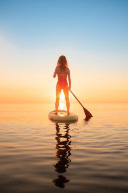 Genç bir kadın yaz tatilinde deniz kenarında ayağa kalkıp kürek çekiyor. Deniz ve gökyüzü arka planında kürek çeken güzel bir kız.