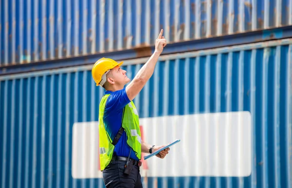 Factory worker man in hard hat holding clipboard checklist and pointing to the sky at container cargo