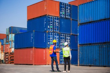 Yelekli kadın ustabaşı dizüstü bilgisayar tutuyor ve kasklı işçi kargo konteynırlarını kontrol ediyor.