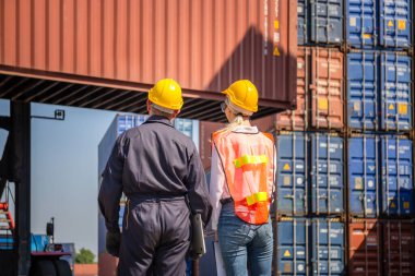 Kask ve güvenlik yeleği giyen işçi elinde dizüstü bilgisayar ve kadın mühendis kontrol konteynerlerini kargo kutusundan yüklüyor.