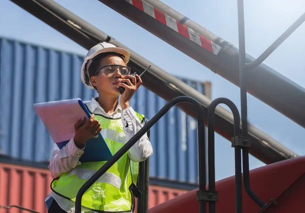 Woman Foreman in hardhat and safety vest holding holding clipboard checklist and talks on two-way radio control loading containers box from cargo