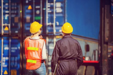 Kask ve güvenlik yeleği giyen işçi elinde dizüstü bilgisayar ve kadın mühendis kontrol konteynerlerini kargo kutusundan yüklüyor.