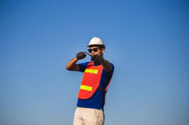 Çelik yelek giyen fabrika işçisi iki yönlü telsiz tutuyor ve önünü işaret ediyor.