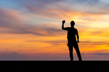 Fabrika işçisinin silueti, zafer kutlamalarıyla birlikte kasket takan genç adam, günbatımı arka planında el ele kutluyor.