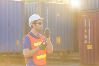 Kask ve güvenlik yeleği giyen ustabaşı iki yönlü telsizle konuşuyor, fabrika işçi kontrol kargo kutusundan konteynırları yüklüyor.