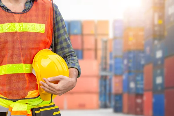 Engineer man in safety vest holding hard hat with blurred cargo container background